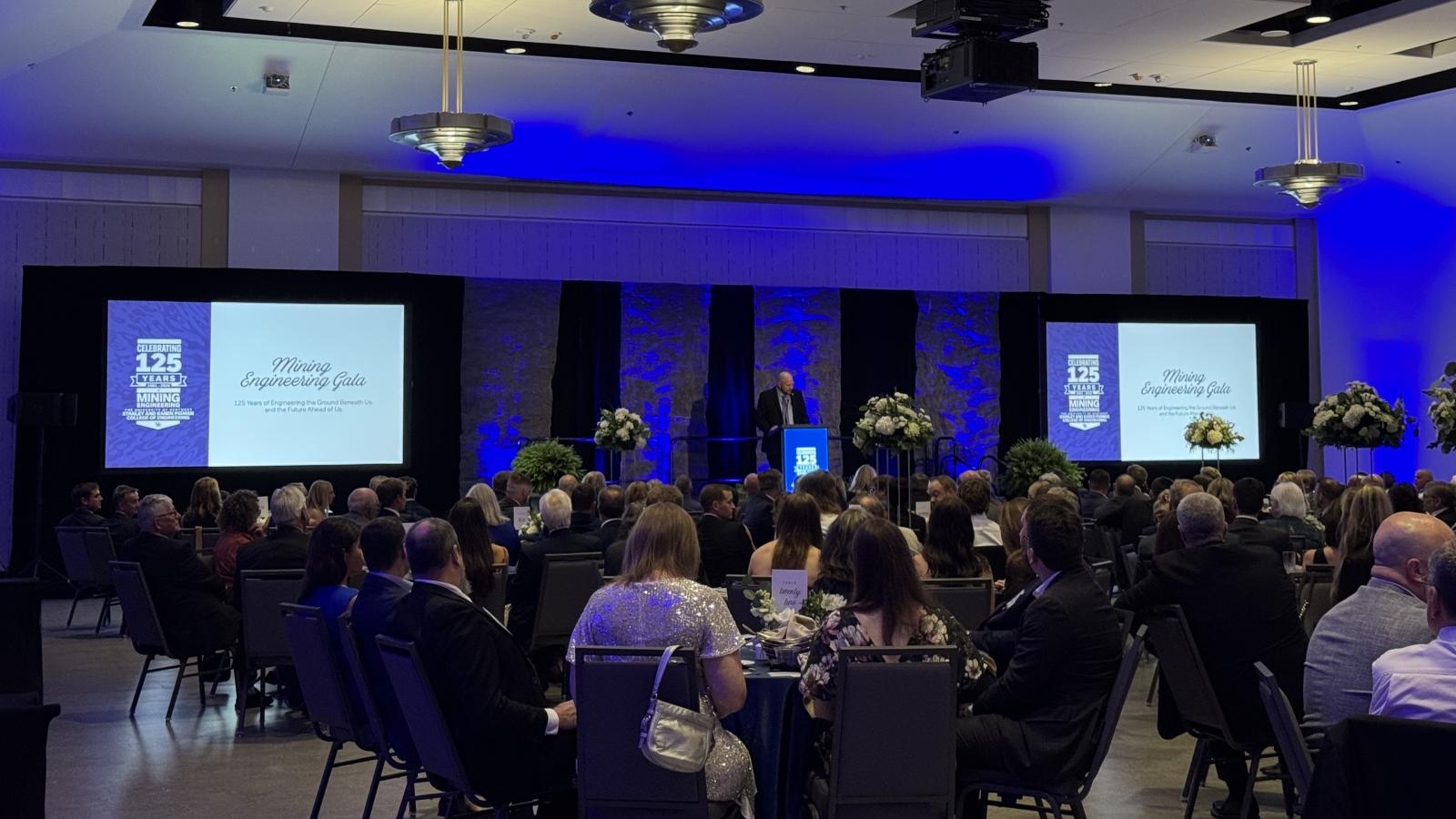 Mining 125th gala room with tables and a speaker at a podium with two screens displaying Mining Engineering Gala