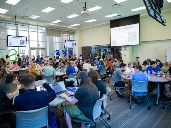 A classroom full of students sitting at tables and a projection screen that says "Engage with an engineer" at the front of the classroom.